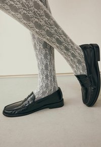 Čierne kožené loafers s lesklým povrchom a dekoratívnym remienkom, doplnené o šedé pančuchy so čipkovaným vzorom, ktoré zdôrazňujú zložitú textúru.
