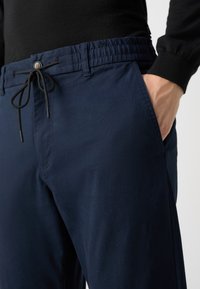 Navy blue pants made of soft fabric with an elastic waistband, button detail, and drawstring. Features side pockets and a smooth texture.