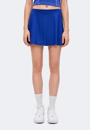 Vrouw draagt een blauwe sportrok, witte sokken en witte sneakers, staat met handen ontspannen langs haar zij op een effen achtergrond.