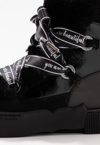 Musta lakattu nahkakenkä, jossa on pörröisiä mustia yksityiskohtia, koristeelliset satiininyörit, joissa on valkoista tekstiä. Paksu musta pohja, jossa on teksturoitu pinta.