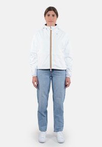 Giacca bianca leggera con cappuccio e dettagli della cerniera gialla, abbinata a jeans azzurri chiari e sneakers bianche, in piedi contro uno sfondo semplice.