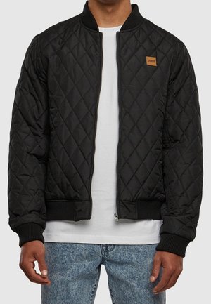 Man wearing a black quilted bomber jacket over a white t-shirt and blue jeans, standing with hands relaxed at sides.