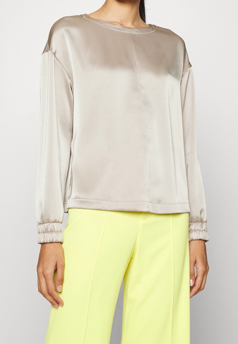 Femme portant un chemisier ample en satin beige à manches longues avec poignets froncés et un pantalon large jaune vif.