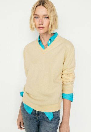 Donna con capelli biondi corti indossa un maglione beige sopra una camicia con colletto blu brillante e jeans blu, in piedi davanti a uno sfondo semplice.