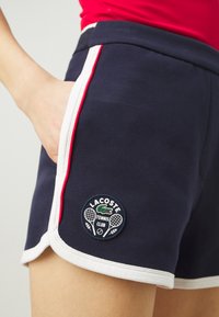 Marineblå shorts laget av teksturert stoff med hvite og røde sideakenter. Har et sirkulært Lacoste Tennis Club-merke på kanten.