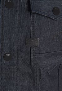 Mörk denimjacka med en texturerad yta, med svarta metallknappar, en bröstficka och en G-Star Originals-logotyp på framsidan.