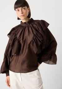 Brun satinbluse med flæset ærmer og høj halsudskæring. Har tekstureret stof, lagdelte flæser og en samlet talje med elastiske manchetter.
