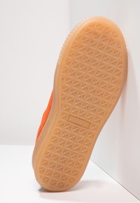Sneaker en daim orange montrant une semelle extérieure en caoutchouc texturé avec un motif en étoile et une marque visible sur le côté.