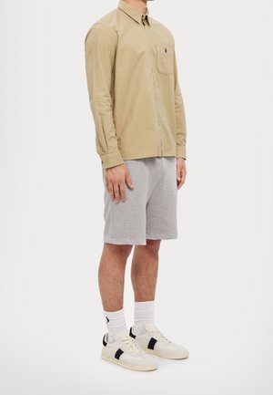 Mann trägt beige langärmeliges Hemd mit Knöpfen, graue knielange Shorts, weiße Socken und weiße Sneakers mit schwarzem Streifen, steht seitlich.