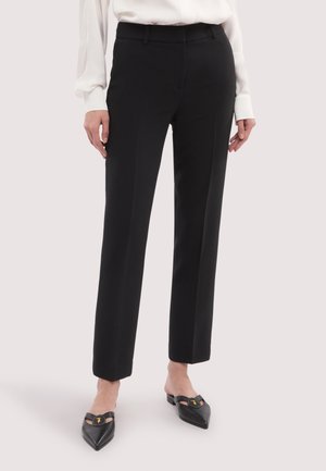 Vrouw die zwarte, op maat gemaakte rechte broek draagt, met een witte blouse met lange mouwen en zwarte platte schoenen met spitse neus en gespsluiting.