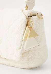 Struktureret cremefarvet stofpose med quiltet design. Har guldhardware og trekantede lædermærker, der tilføjer detaljer og kontrast.