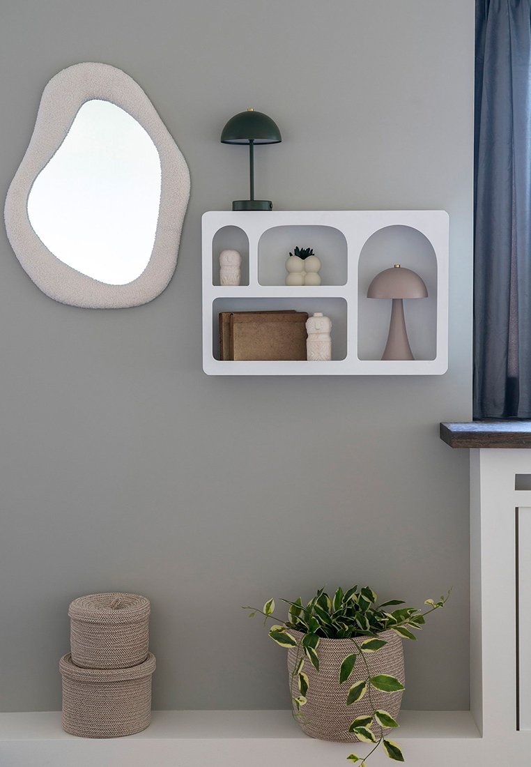 Mensola bianca a muro con scomparti arrotondati, contenente una lampada, figure decorative e libri. Include uno specchio rotondo testurizzato e piante in vasi intrecciati.