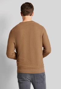 Brauner strukturierter Pullover mit Rundhalsausschnitt und langen Ärmeln, ausgestattet mit einem gerippten Saum. Das Modell trägt graue Hosen.