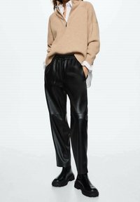 Beżowy sweter z zamkiem błyskawicznym o fakturze prążkowanej, zestawiony z czarnymi spodniami z ekologicznej skóry o wysokim stanie i masywnymi czarnymi butami.