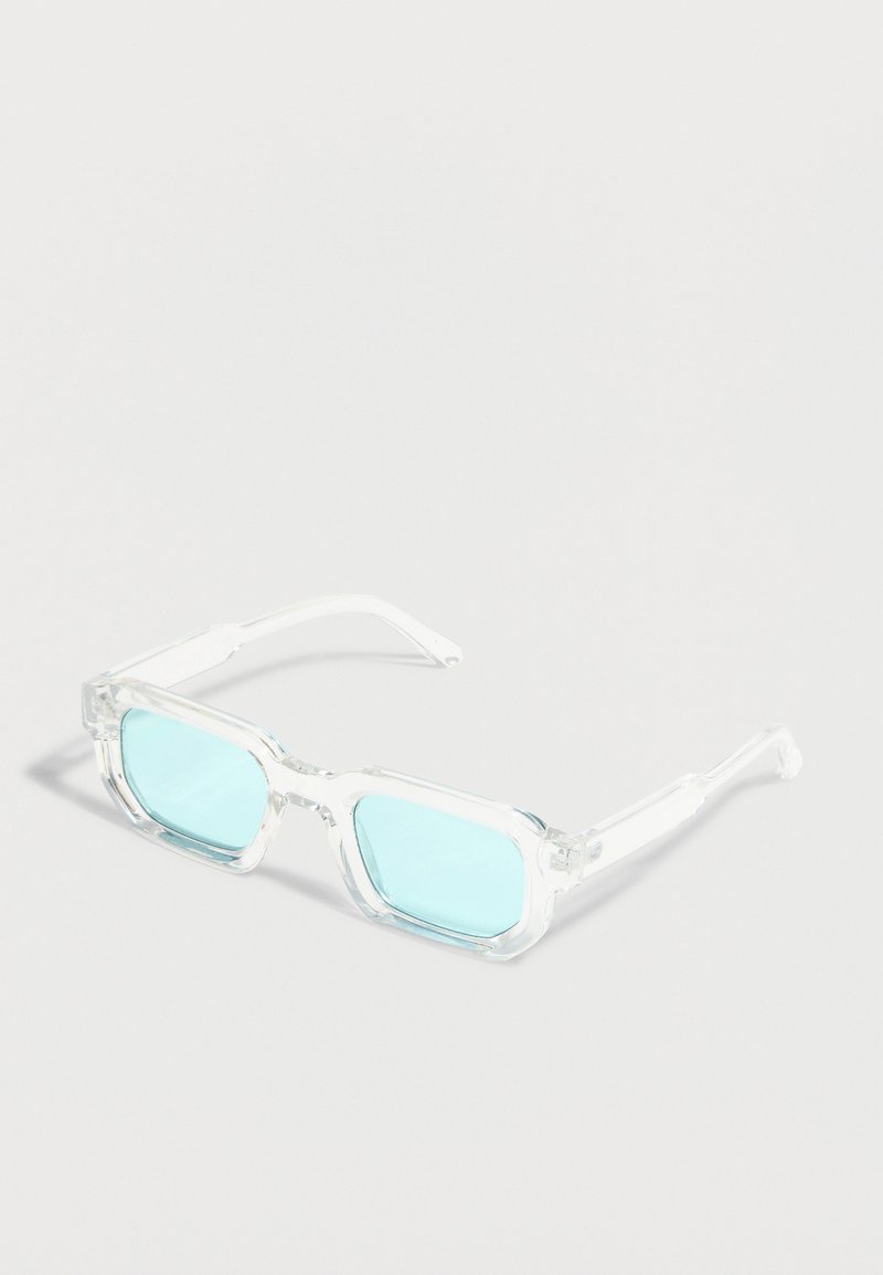 Lunettes rectangulaires transparentes avec des montures épaisses transparentes et des verres légèrement teintés en bleu clair reposant sur une surface blanche.