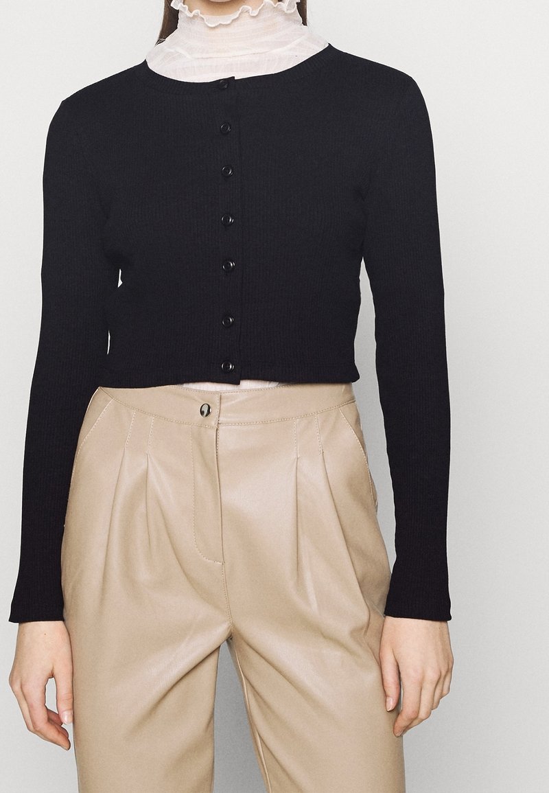 Femme portant un cardigan court noir boutonné sur un col roulé blanc à volants et un pantalon plissé beige taille haute.