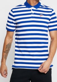 Blå och vitrandig polotröja med korta ärmar, treknappsknäppning och en liten röd logotyp. Tyget är en mjuk bomullsblandning.