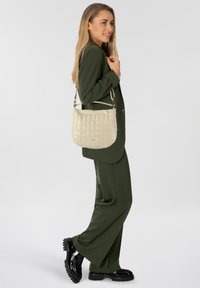 Borsa beige in ecopelle con motivo a coccodrillo testurizzato, forma curva, dimensione media e tracolla regolabile, tenuta da una modella con un completo oliva.