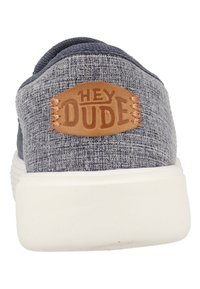 Blauwe stoffen instapschoen met een witte zool, met een bruine leren patch met het label "HEY DUDE" en een gestructureerd oppervlak op de bovenkant.
