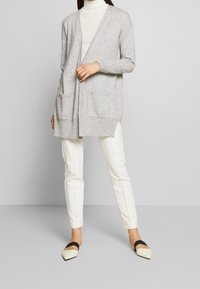 Ljust grå lång cardigan med öppen framsida, två fickor fram och ribbade manschetter, tillsammans med vita smala byxor och spetsiga svart-vita ballerinas.