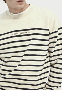 Camisa de manga larga color crema con rayas horizontales azul marino, cuello redondeado y detalle del logo en el pecho. Tejido suave y diseño de corte relajado.