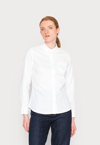Camicia bianca con maniche lunghe e colletto, caratterizzata da una tasca sul petto e un orlo dritto. Indossata con jeans in denim scuro.