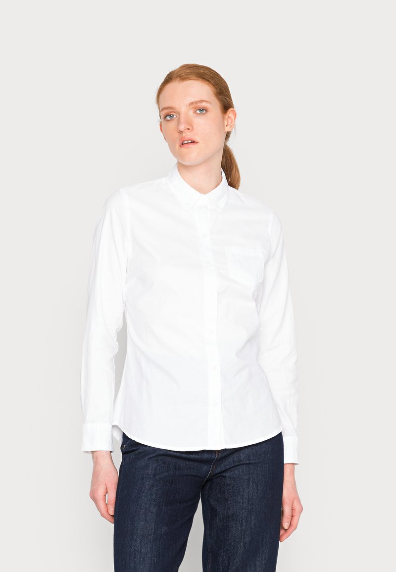 Camicia bianca con maniche lunghe e colletto, caratterizzata da una tasca sul petto e un orlo dritto. Indossata con jeans in denim scuro.