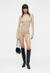 Μπεζ ολόσωμο κορμάκι με μακριά μανίκια και μαύρο μοτίβο σε σχήμα ημισελήνου, συνδυασμένο με ψηλές μαύρες δερμάτινες μπότες και μικρή μαύρη τσάντα.