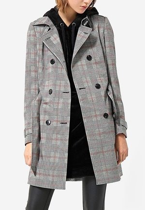 Femme portant un manteau croisé gris à carreaux par-dessus une robe à capuche noire et un pantalon en cuir noir, debout sur un fond blanc.