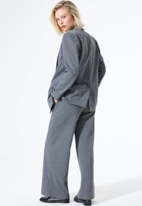 Costume  à rayures gris avec une veste ajustée et un pantalon large. Tissu lisse, éclat subtil et coupe décontractée. Des chaussures noires complètent le look.