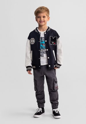 Młody chłopiec w czarno-białej kurtce varsity, koszulce z grafiką dinozaura, szarych spodniach cargo i czarnych trampkach, stojący i uśmiechający się.