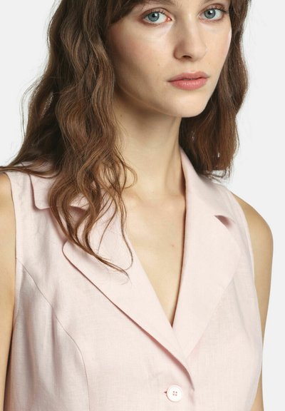 Žena s vlnitými hnedými vlasmi, oblečená v bezrukávovom svetloružovom saku s hlbokým výstrihom do V na bielom pozadí.