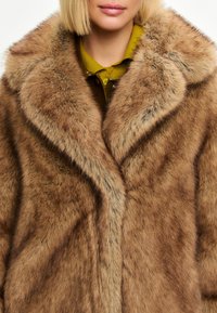 Faux fur frakke i brunt med en blød tekstur, der har en bred krave og slag. Under den ses en grøn bluse med krave.