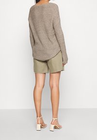 Beige gestrickter Pullover mit lockerer Passform, rundem Ausschnitt und gerippter Textur, kombiniert mit olivgrünen Shorts und weißen Blockabsatzsandalen.