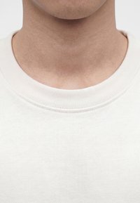 Nahaufnahme des Halses und unteren Gesichtsbereichs einer Person, die ein schlichtes weißes T-Shirt mit Rundhalsausschnitt vor einfachem Hintergrund trägt.