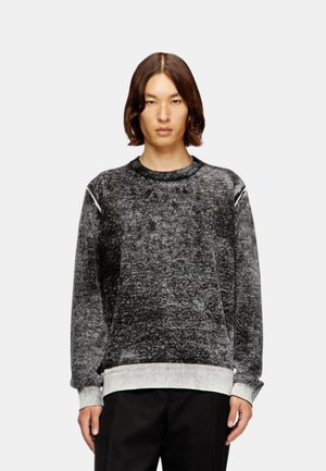 Personne aux cheveux bruns mi-longs portant un pull texturé noir et gris à manches longues et un pantalon noir, debout devant un fond uni.