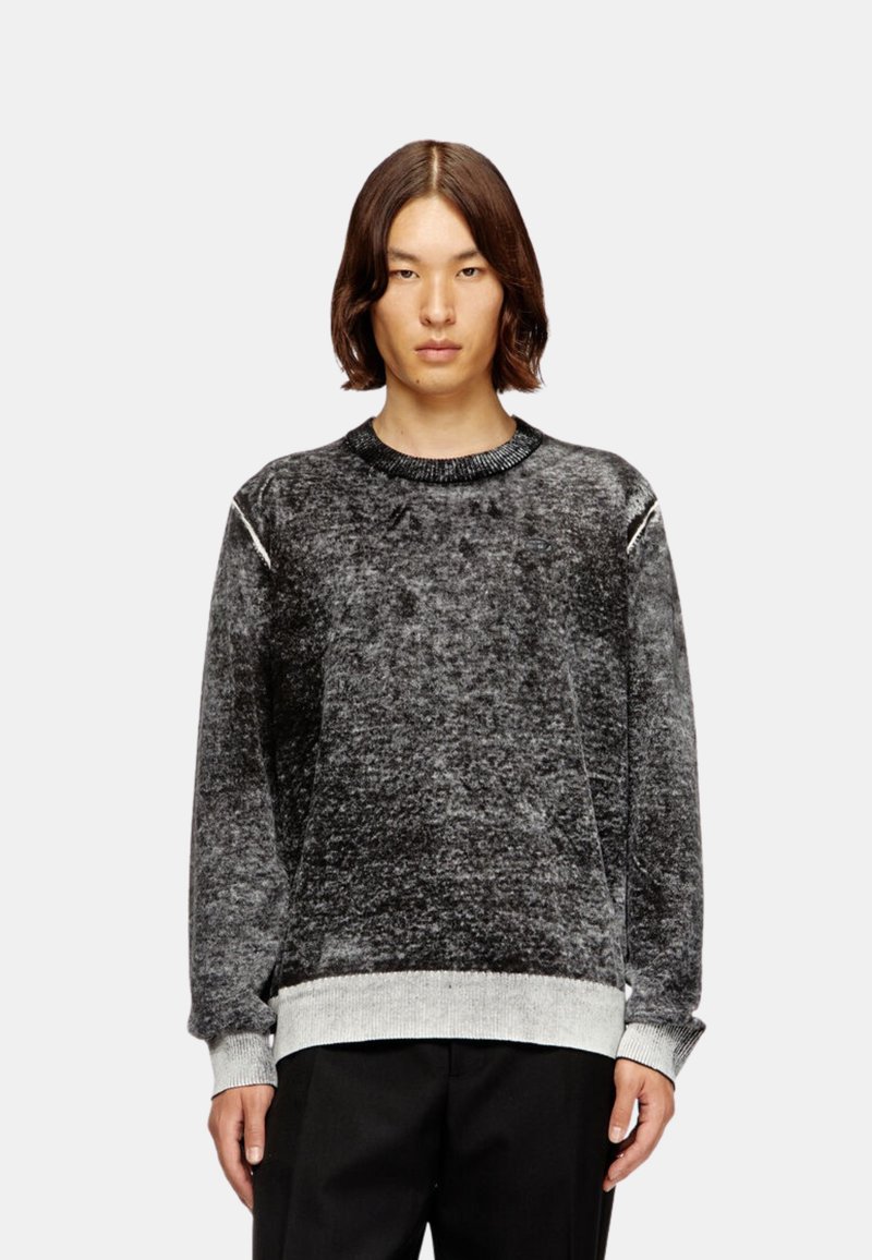 Personne aux cheveux bruns mi-longs portant un pull texturé noir et gris à manches longues et un pantalon noir, debout devant un fond uni.