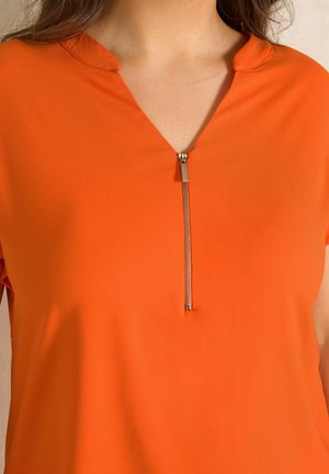 Kobieta ubrana w jasnorudą koszulkę z krótkim rękawem i srebrnym półzamkiem przy dekolcie.