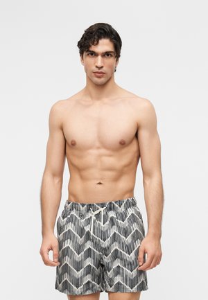 Άνδρας χωρίς πουκάμισο με σγουρά σκούρα μαλλιά που φοράει μαύρα και λευκά μαγιό με ζιγκ-ζαγκ σχέδιο, στέκεται μπροστά σε απλό ανοιχτό φόντο.