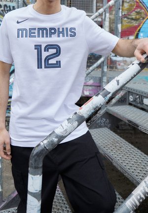 Person mit weißem Memphis 12 Sportshirt und schwarzer Hose, der ein mit Graffiti bedecktes Metallgeländer an einer Außentreppe festhält.