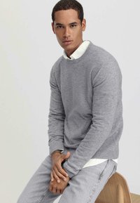 Grauer gestrickter Pullover mit runden Ausschnitt, über einem weißen Hemd mit Kragen getragen. Hellgraue Jeans, auf einem Holzstuhl sitzend. Glatte Textur.
