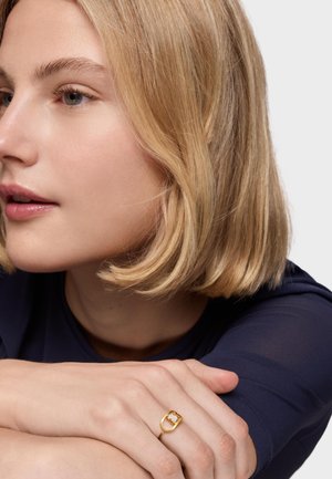 Kobieta z krótkimi blond włosami opierająca podbródek na skrzyżowanych ramionach, nosząca złoty pierścionek z białym i złotym detalem na serdecznym palcu lewej ręki.