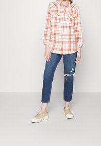 Camisa de cuadros naranja y blanca con botones y dos bolsillos en el pecho, combinada con jeans azules desgastados y zapatillas beige. Ajuste casual y relajado.