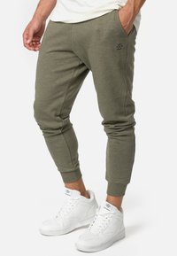 Olijfgroene joggingsbroek in een zachte stof met een taps toelopende pasvorm, voorzien van zijzakken en een geborduurd logo nabij de taille. Witte sneakers gedragen.