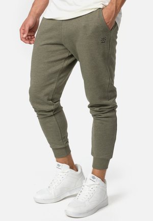 Olive green jogger pants in a soft fabric with a tapered fit, featuring side pockets and an embroidered logo near the waist. White sneakers worn.