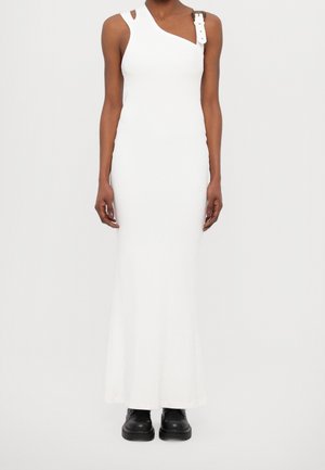 Robe longue blanche sans manches avec un décolleté asymétrique et un détail de bride avec boucle, portée avec des bottes noires épaisses.