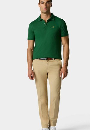 Zielona koszulka polo z krótkim rękawem i kołnierzykiem, zestawiona z beżowymi spodniami i białymi sneakersami. Koszulka ma małe logo na klatce piersiowej.