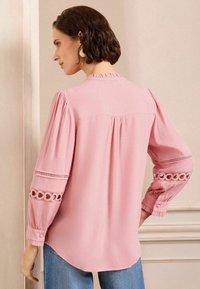 Blusa rosa con lunghe maniche puff caratterizzata da pattern a intaglio e delicate rifiniture, dettagli a pieghe sul retro e orlo arrotondato.