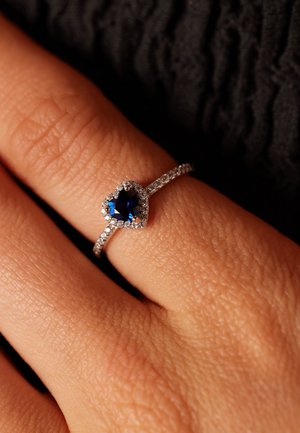 Zilveren ring met een hartvormige blauwe edelsteen in het midden, omringd door kleinere heldere stenen op een textuur band, geplaatst op een hand.