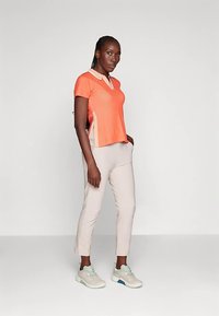 Pomarańczowa koszulka polo z krótkim rękawem, dekoltem w serek i bocznymi rozcięciami, zestawiona z jasnobeżowymi dopasowanymi spodniami i szarymi trampkami.